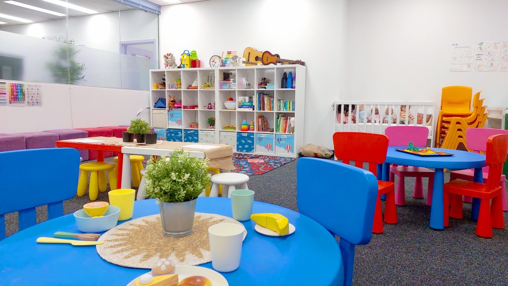 Photo of Room 3-4, the childcare playroom, on Level 3, Berkeley Business Institute (BBI) and Wellington Education Group (WEG) Sydney CBD City campus. The angle shows the inside of the classroom, and the kids toy tables and chairs, a crib with toys and dolls, and the shelves along the classroom wall.