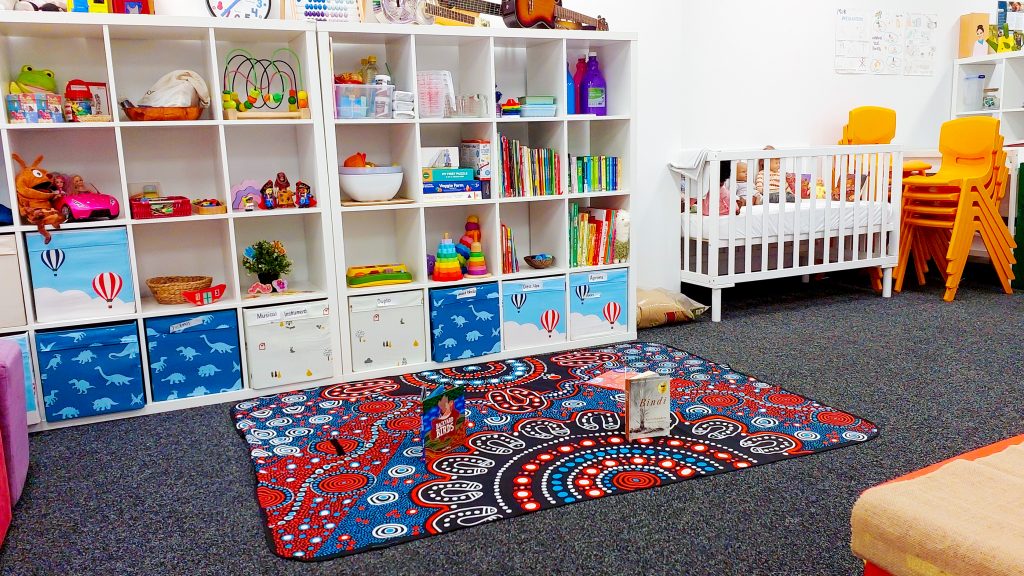 Photo of Room 3-4, the childcare playroom, on Level 3, Berkeley Business Institute (BBI) and Wellington Education Group (WEG) Sydney CBD City campus. The angle shows the shelves along the classroom wall, filled with toys and books, a crib filled with toys and dolls, and a large play mat or rug with a book on it.